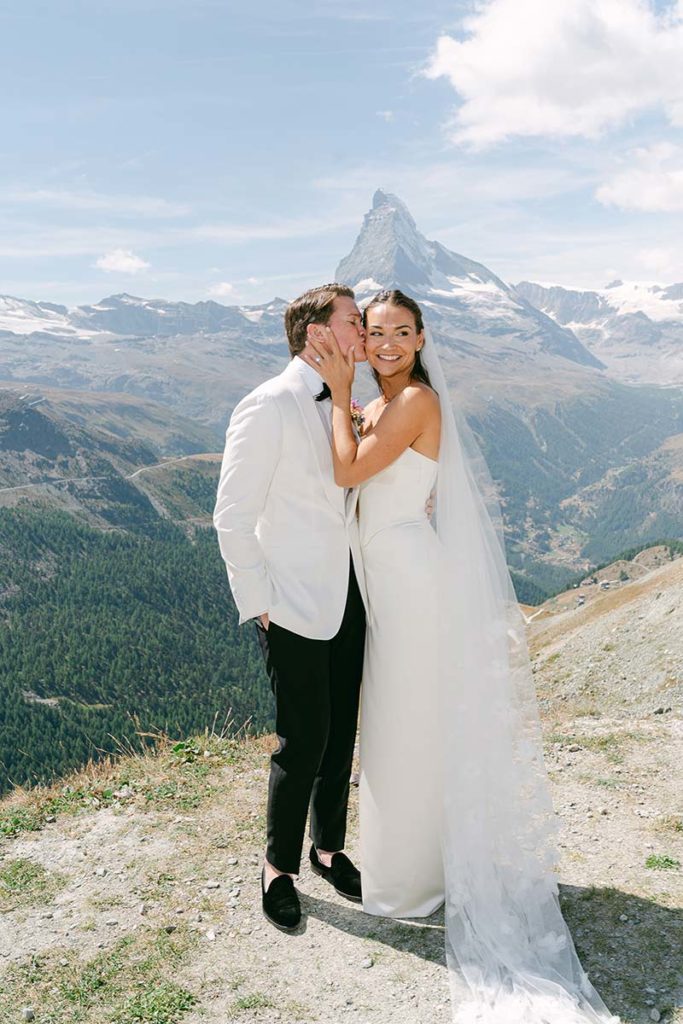 A bride and groom pose outdoors on a mountain slope in Switzerland with the Matterhorn in the background; the groom kisses the bride's cheek as she smiles, capturing a perfect moment for their Zermatt wedding videography.