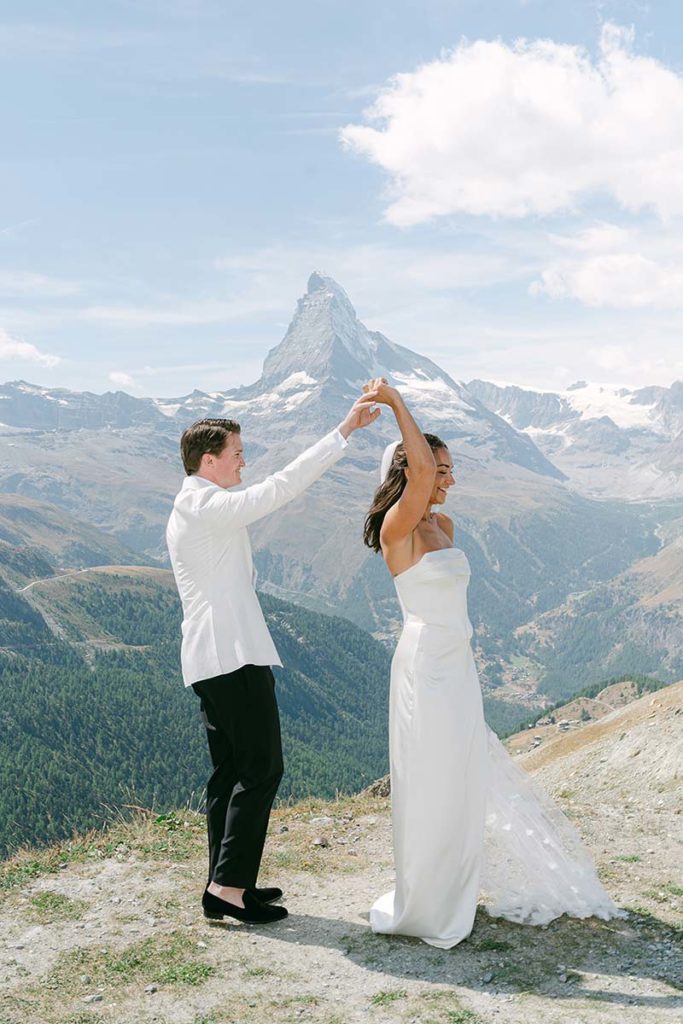 A couple in wedding attire dances outdoors on a mountain, with the Matterhorn peak visible in the background under a partly cloudy sky during their Wedding in Zermatt