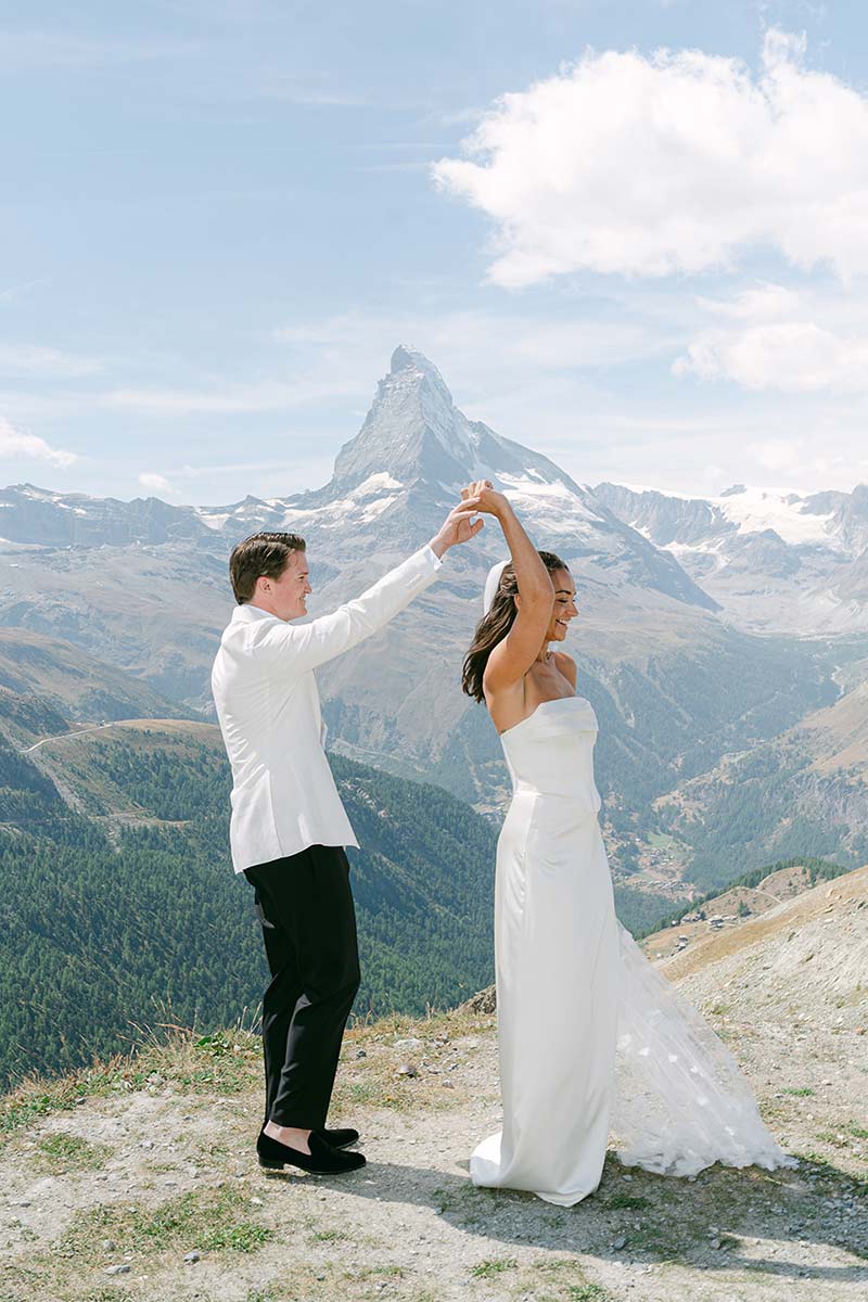 Ein Paar in Hochzeitskleidung tanzt draußen auf einem Berg, mit dem Matterhorn im Hintergrund unter einem teilweise bewölkten Himmel während ihrer Hochzeit in Zermatt.
