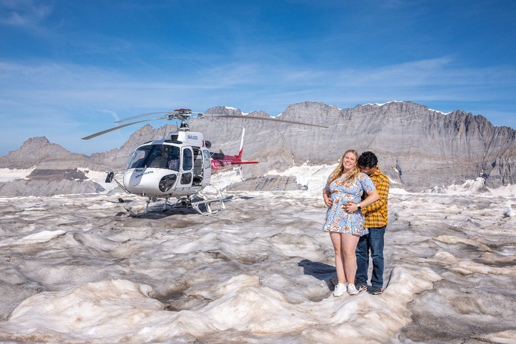 A couple smile and embrace standing on a glacier with a helicopter behind them photographed during their Swiss Proposal 