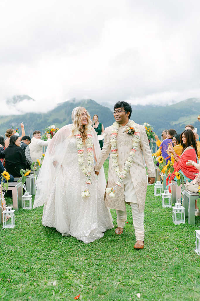 A bride and groom walk back down the aisle during their wedding celebration in the Swiss Alps. The bride wears a wedding dress and the groom wears traditional Indian attire. Both are smiling and wearing flower garlands