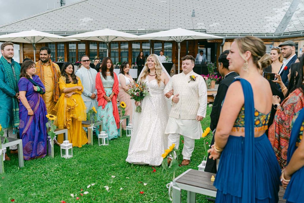 A bride walks down the aisle with her brother during her Swiss Alpine Wedding celebration. The guests are dressed in brightly colour Saris