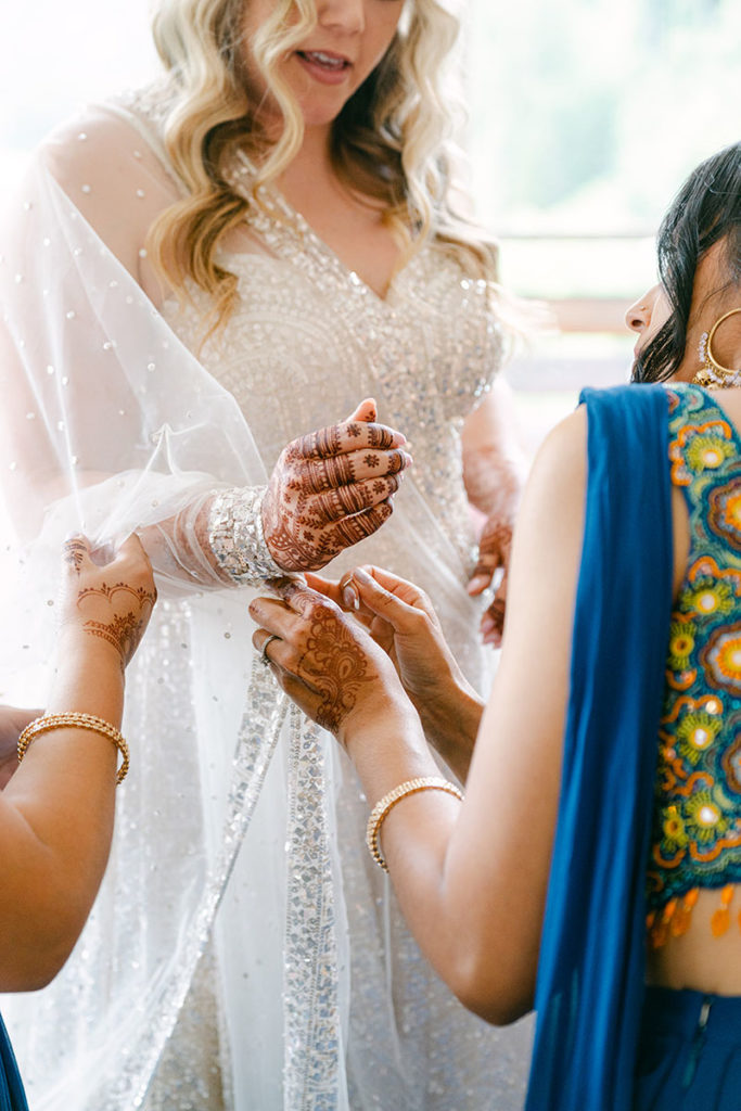 A bride stands in her wedding gown. She has henna patterns on her hands and her wedding party help adjust her veil during her Swiss Alpine wedding celebration