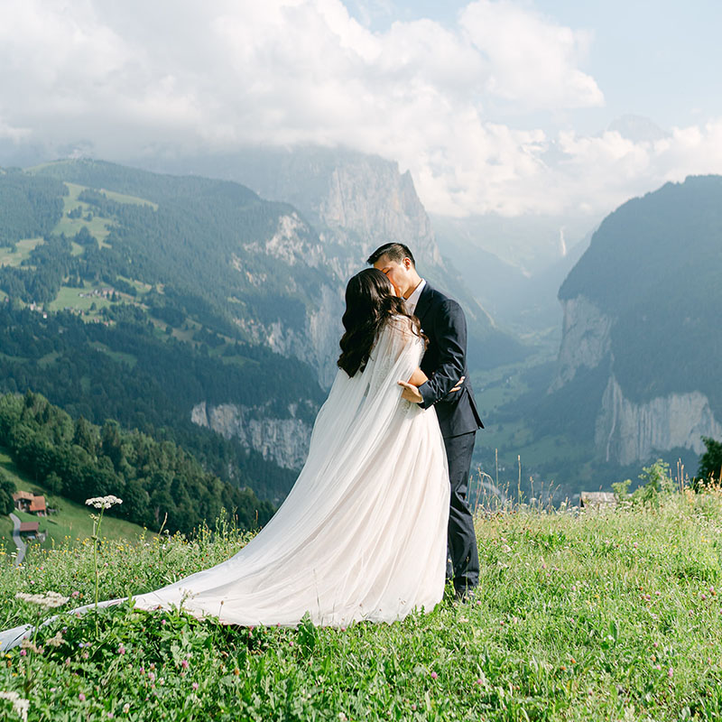 A bride and groom kiss on a grassy hilltop in Switzerland, with majestic mountains and a cloudy sky in the background—perfect for stunning wedding videography.