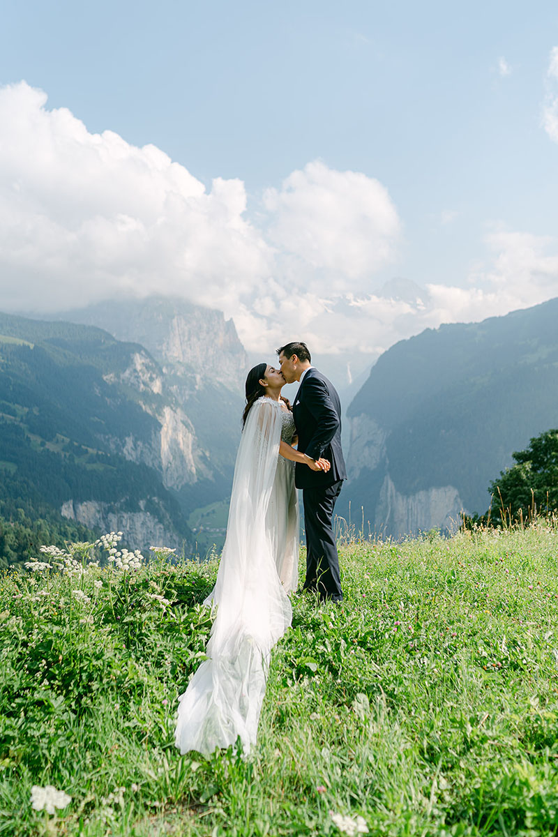 A bride and groom kiss on a grassy hilltop with mountains and clouds in the background, perfectly captured by an experienced elopement videographer, Story Of Your Day