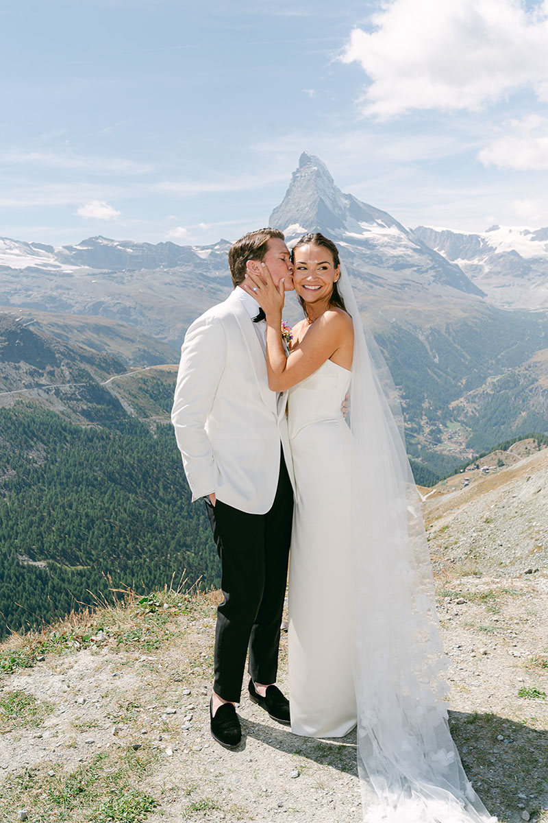 Bride and groom pose outdoors in wedding attire, smiling with mountains and the Matterhorn in Switzerland behind them under a sunny sky