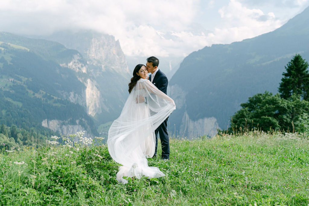 A bride and groom stand on a grassy hillside, embracing with mountains and a cloudy sky in the background. The bride’s veil flows behind her during their Wengen Wedding