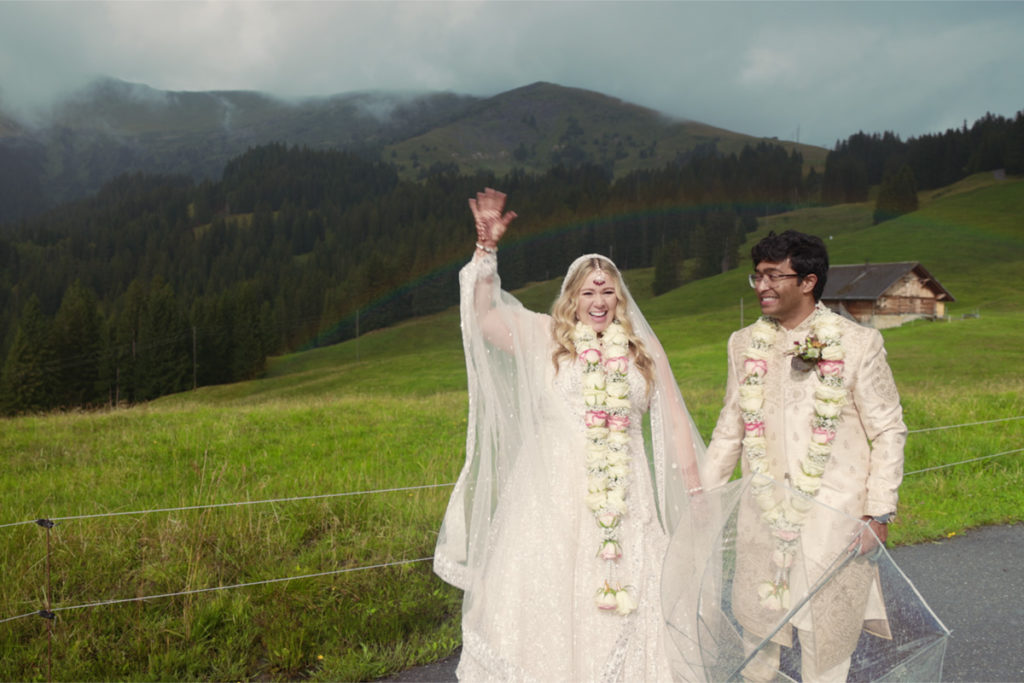 A bride and groom wearing traditional attire and floral garlands stand on a rural roadside, holding hands and smiling, with green hills and mountains in the background.