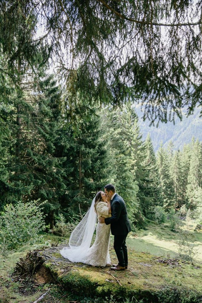 A bride and groom stand on a mossy rock, kissing in a forest clearing with tall evergreens and mountains behind them—an intimate moment captured for Swiss elopement photography.