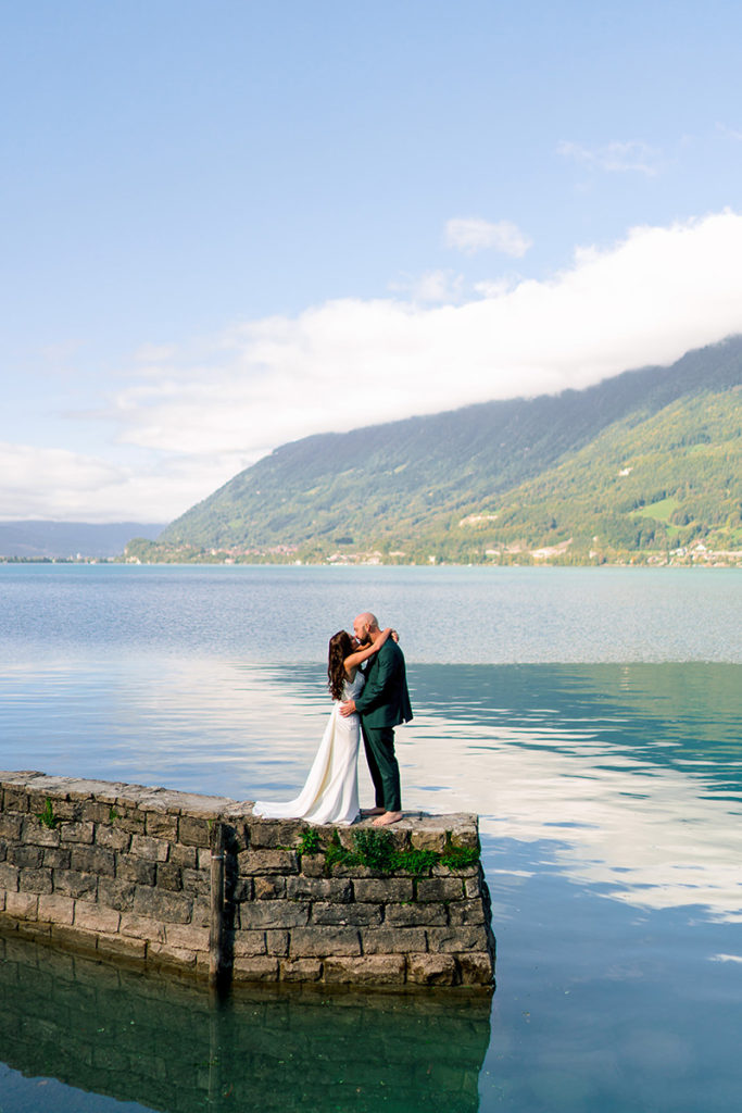A couple in wedding attire stands and embraces on a stone pier overlooking a calm lake with Switzerland’s mountains in the background, capturing a romantic elopement moment.