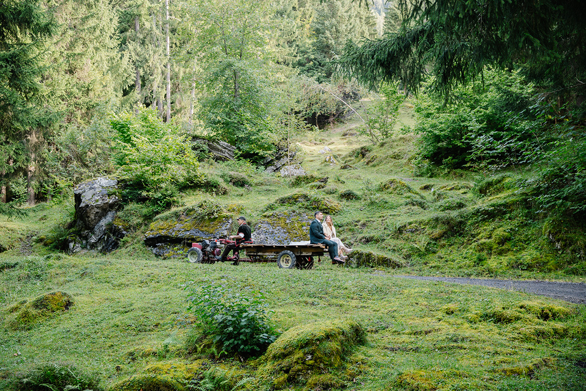 A couple sit on a wooden cart in a green, mossy forest clearing surrounded by trees, rocks, and grass—perfect for an adventurous Switzerland elopement, captured beautifully by Story Of Your Day