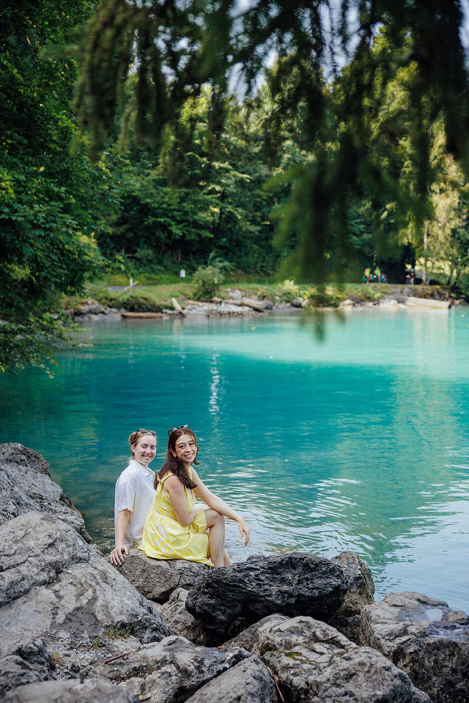 Two people sit on large rocks by a turquoise lake surrounded by lush greenery, smiling toward the camera—an idyllic moment perfect for an elopement photography portfolio.