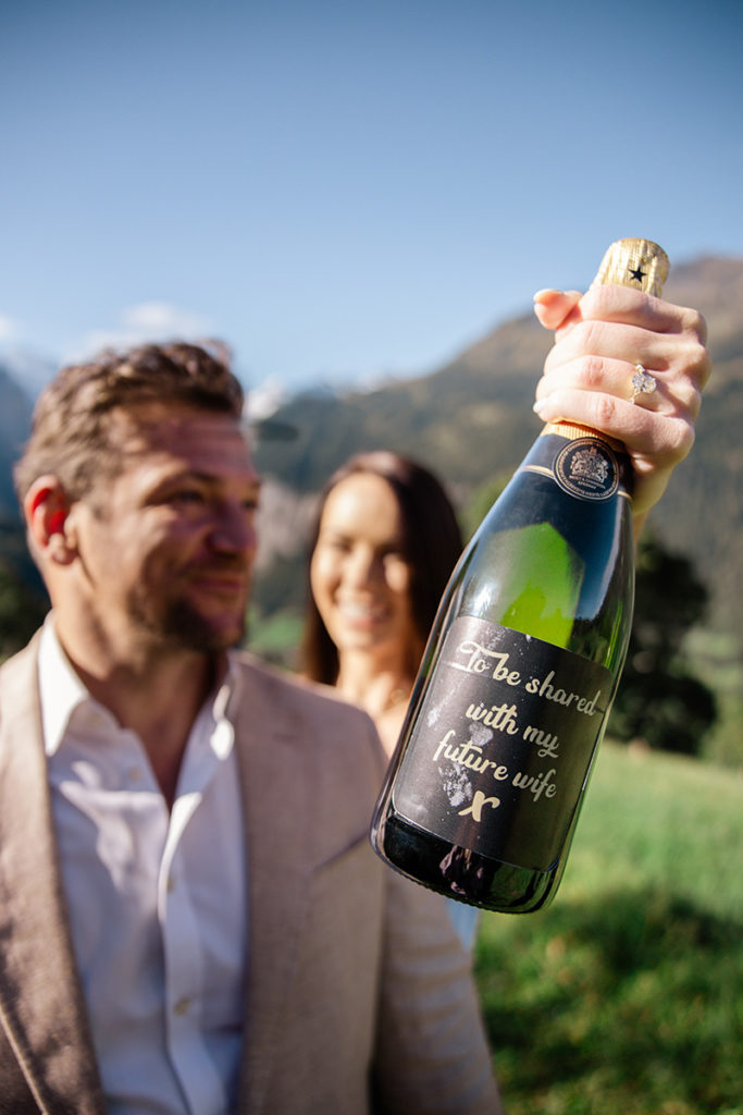 A woman holds a champagne bottle labeled "To be shared with my future wife" beside a man, both standing outdoors in a grassy, mountainous in the Swiss Alps. An unforgettable moment perfect for any proposal photography portfolio.