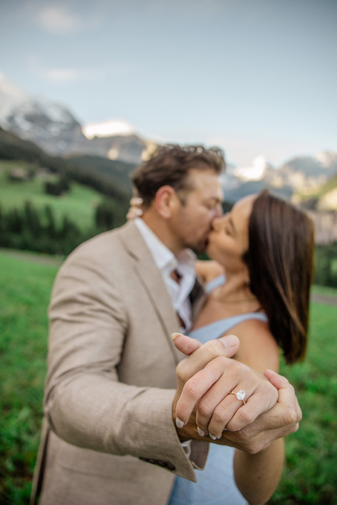 A couple stands in a green field above the Swiss Town of Wengen with mountains behind them, holding hands and kissing. The woman’s hand, highlighted with an engagement ring, captures the essence of proposal photography for your portfolio.