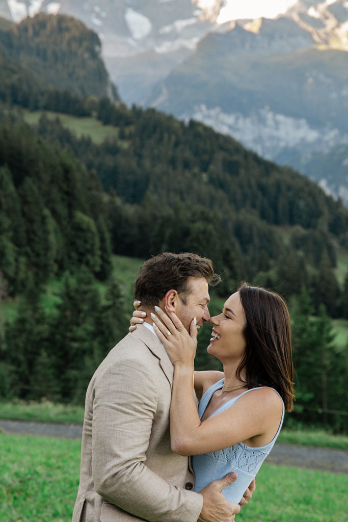 A couple stands embracing and smiling at each other in a grassy area with forested mountains and snow-capped peaks in the background, beautifully capturing a tender moment for their proposal photography portfolio.
