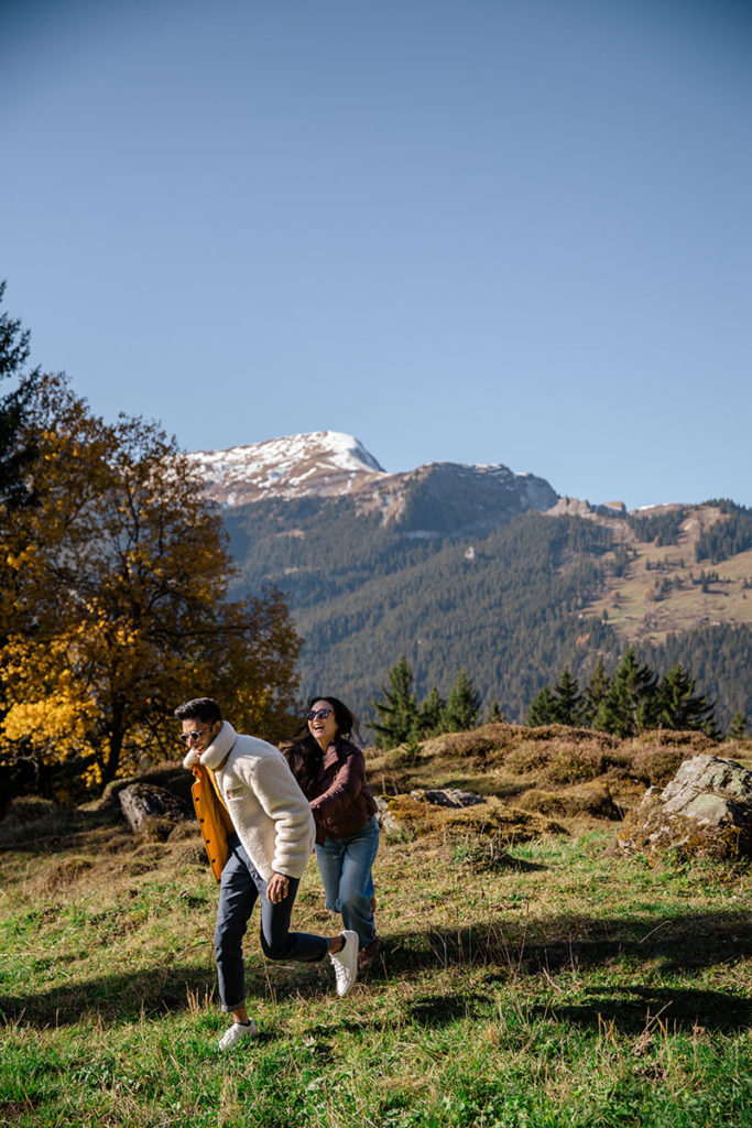 A man and a woman walk together in a grassy, mountainous area with trees and a snow-capped peak in the background under a clear blue sky, setting the perfect scene for a secret proposal.