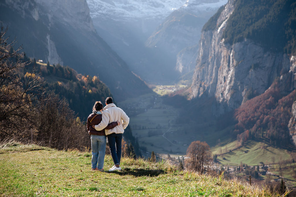 A couple stand on a grassy Wengen meadow with their backs to the camera, overlooking a the Lauterbrunnen valley - an ideal spot for a surprise proposal amid tall cliffs and distant mountains.