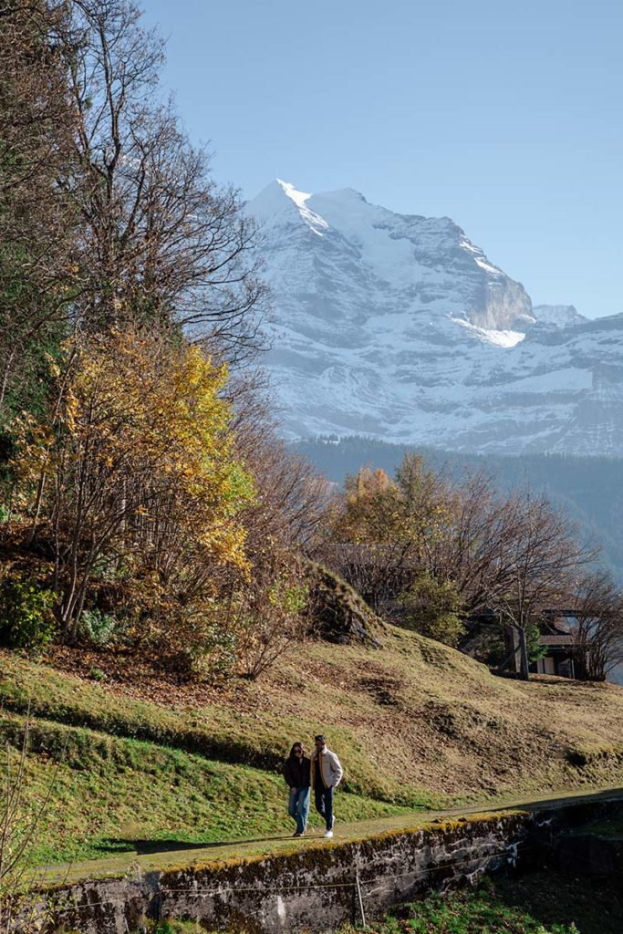 Two people walk along a path surrounded by autumn trees, with a snow-covered mountain in the background under a clear sky—the perfect setting for a secret Wengen proposal.