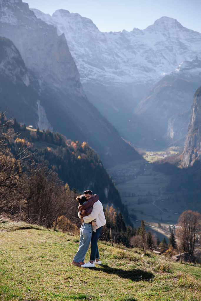 A couple embraces on a grassy hillside above Wengen after a surprise proposal, with a scenic valley and snow-capped mountains of the Jungfrau in the background.