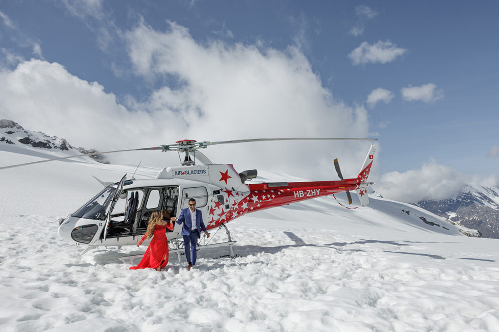 A man and woman in formal attire stand beside a red and white helicopter on a snowy mountain landscape under a partly cloudy sky, creating a striking shot perfect for any proposal photography portfolio.
