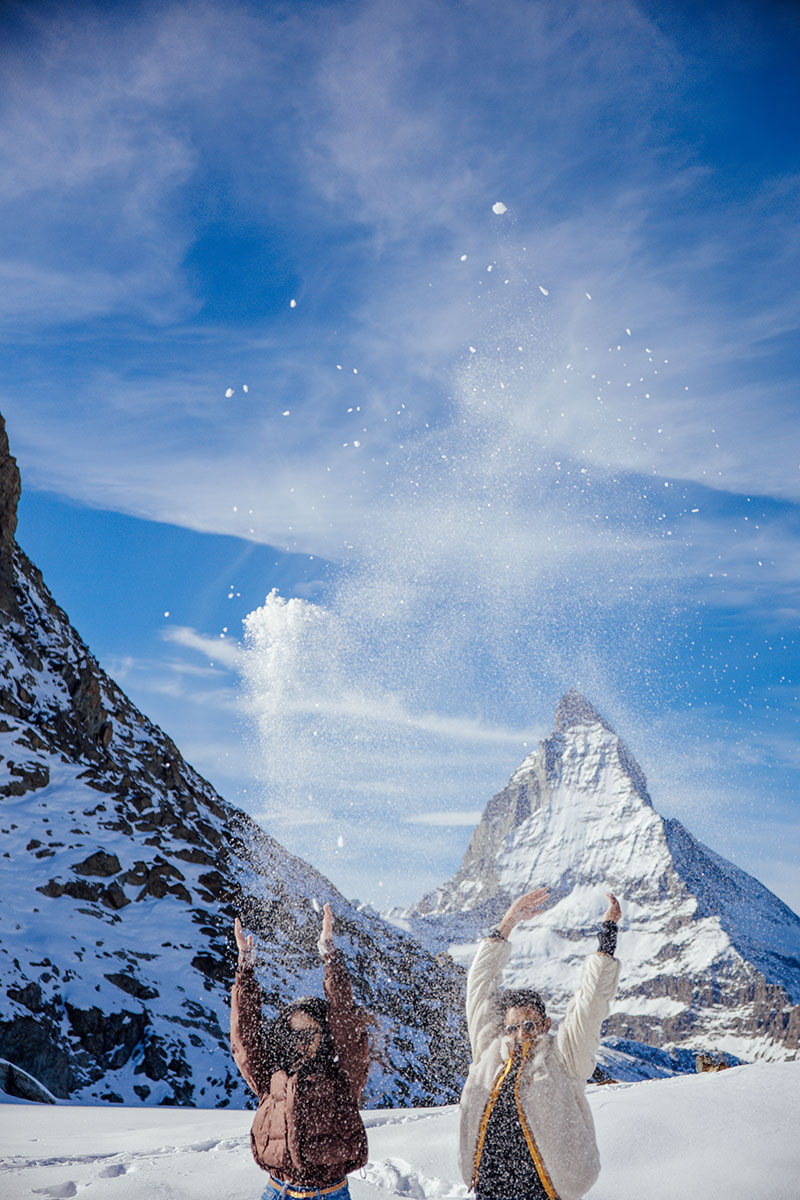 Un couple vêtu de vestes d'hiver jette de la neige en l'air devant le Cervin enneigé sous un ciel bleu lors de leur séance photo de fiançailles à Zermatt avec Story Of Your Day.