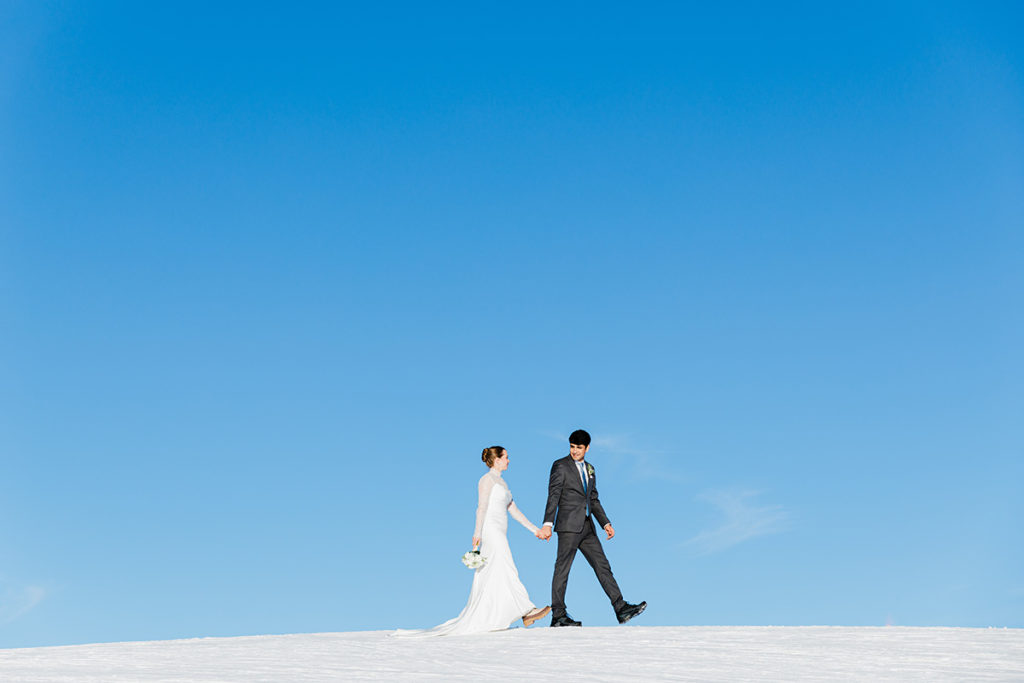 A bride and groom hold hands while walking on snow under a clear blue sky on the top of Männlichen above Wengen during their Swiss Mountain elopement.