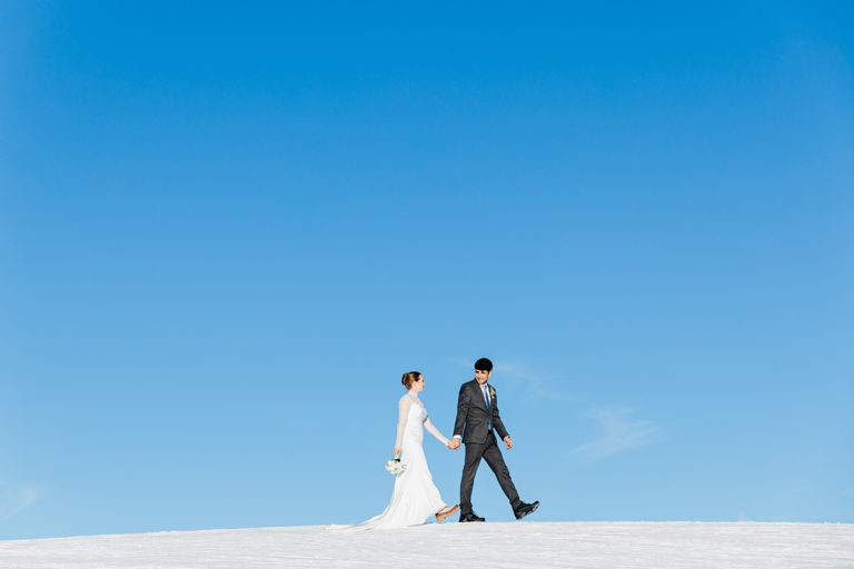 A bride and groom hold hands while walking on snow under a clear blue sky on the top of Männlichen above Wengen during their Swiss Mountain elopement.