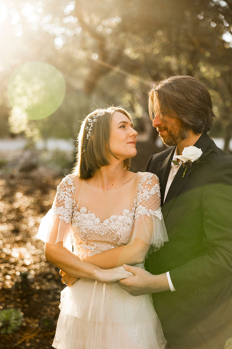A bride and groom stand outdoors in sunlight, embracing and looking at each other during their Los Angeles wedding. the bride wears a lace dress; the groom is in a suit with a boutonniere.