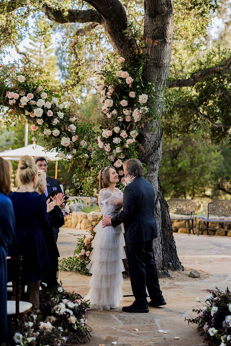 A bride and groom stand facing each other under a large tree decorated with flowers as guests look on, during an outdoor Los Angeles wedding ceremony.