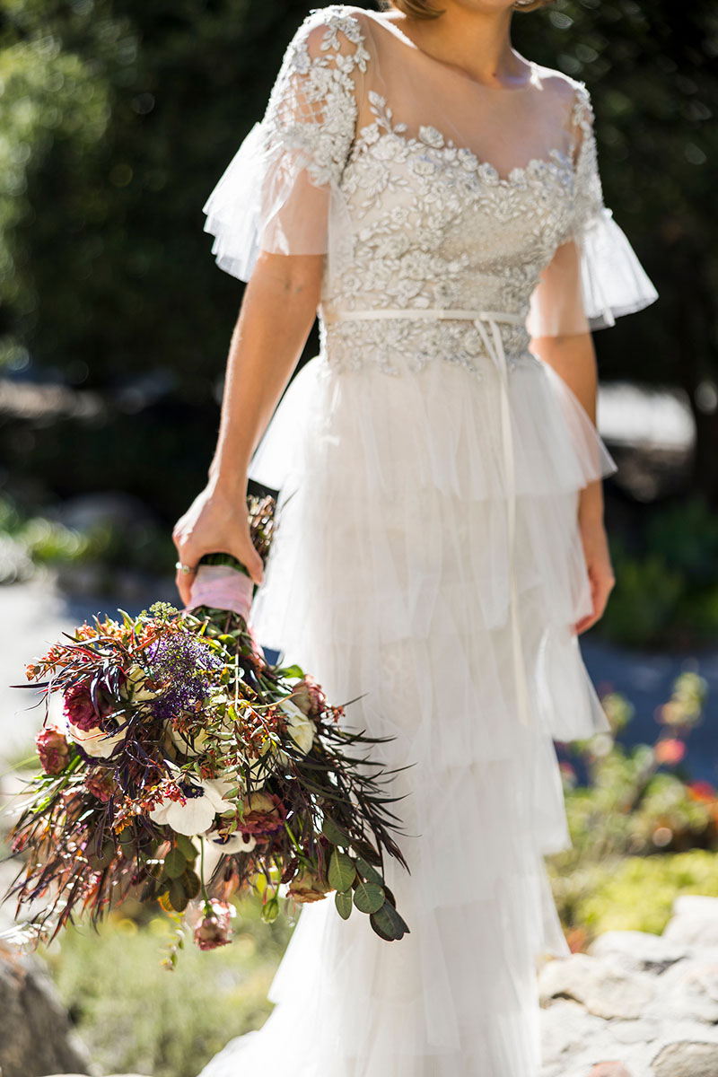 A woman in a white lace wedding dress holds a large bouquet of flowers outdoors, capturing the elegance of this Los Angeles Wedding