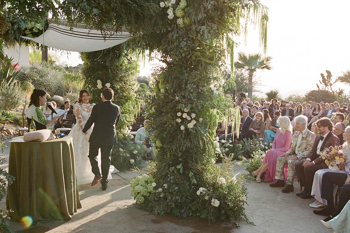 A bride and groom stand under a floral wedding arch during an outdoor Switzerland wedding ceremony as guests watch from their seats.
