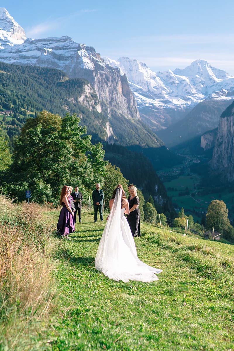 A bride in a white dress and veil walks toward a small outdoor Switzerland wedding ceremony set on a grassy hillside in Wengen with mountains and valleys in the background