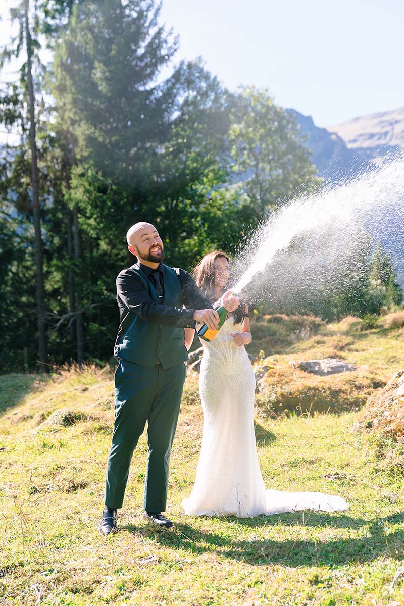 A man in formal attire pops a champagne bottle, spraying foam, while a woman in a white dress stands beside him outdoors in a grassy, mountainous area during their elopement in Wengen