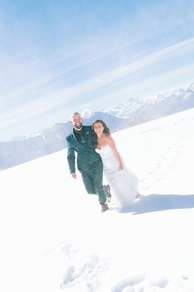 A bride and groom, dressed in wedding attire, run together through deep snow on a mountain in Switzerland, distant peaks and a clear blue sky behind them—an unforgettable scene perfect for Switzerland weddings or elopements.
