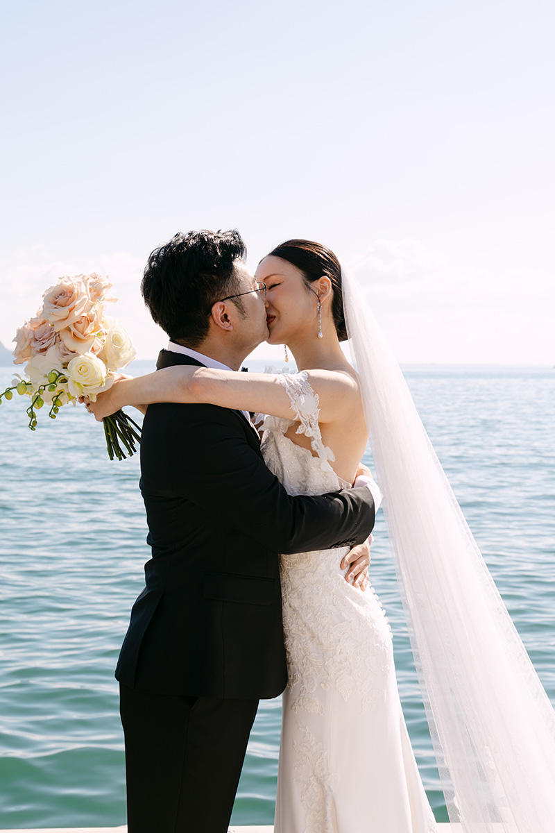 A bride and groom embrace and kiss by the water; the bride holds a bouquet of roses and wears a long veil, capturing the romance of their Isola Del Garda elopement