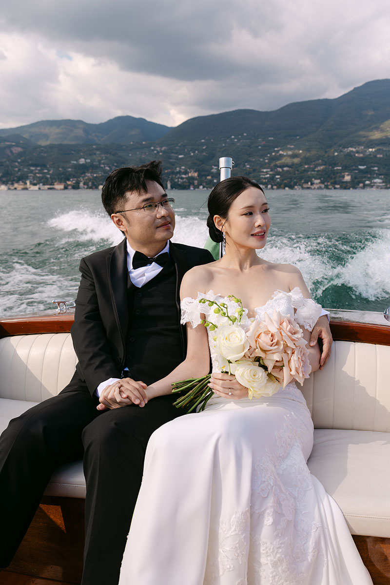 A bride and groom sit together on a boat, holding hands. The bride holds a bouquet of roses as mountains and water set a romantic backdrop during their Isola Del Garda elopement