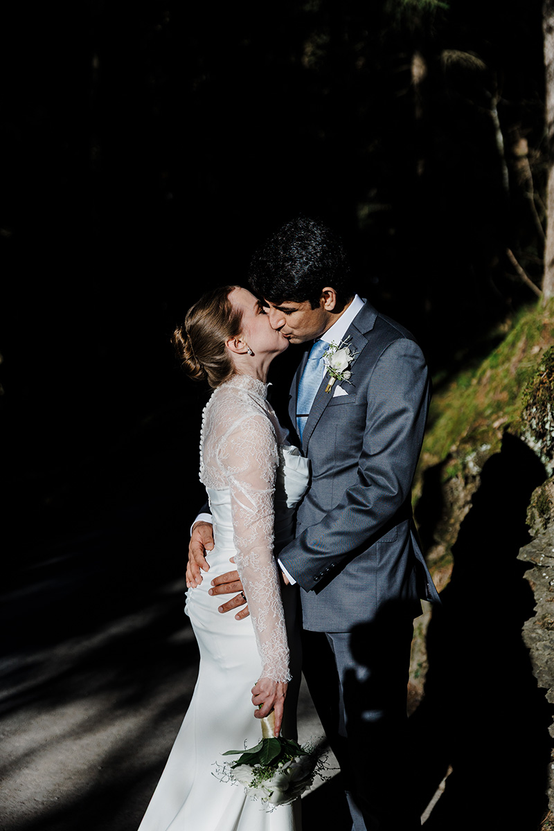 A bride and groom kiss outdoors in formal wedding attire, with the bride holding a small bouquet and sunlight illuminating them against a dark background during their Wengen Elopement