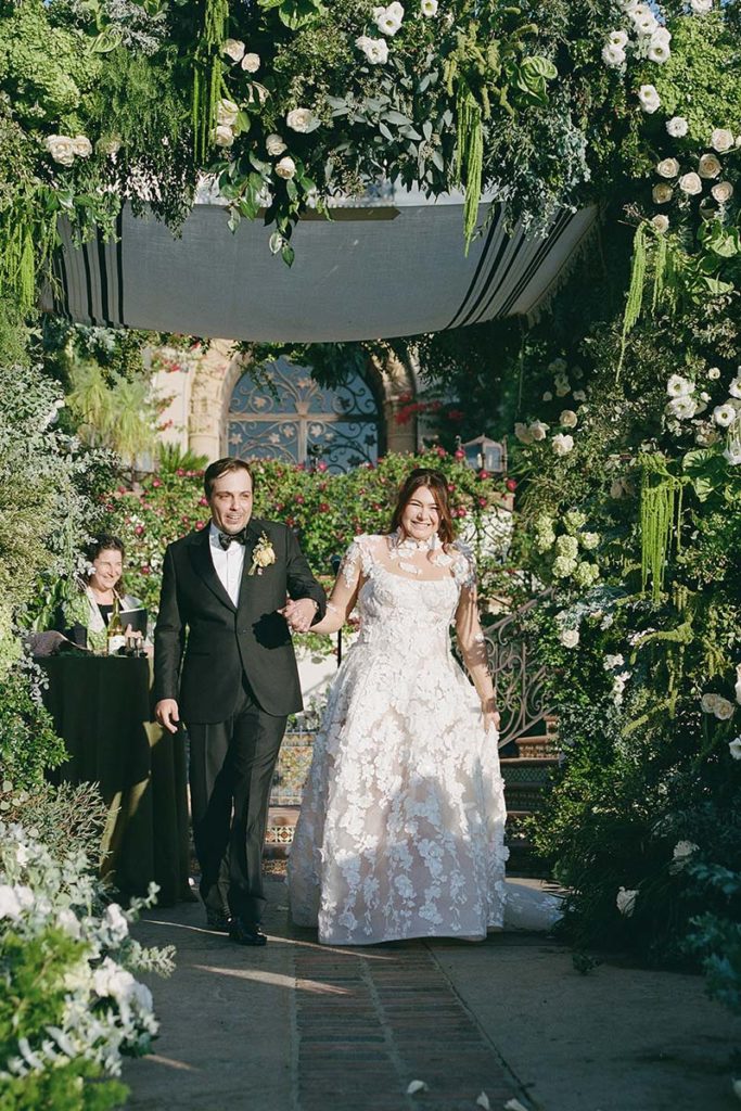 A bride in a white lace gown and a groom in a black tuxedo walk together under a floral arch at an outdoor Switzerland wedding ceremony.