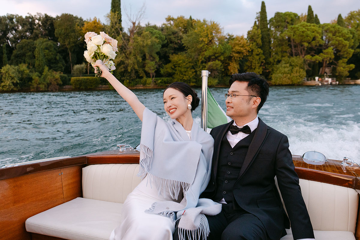 A couple dressed formally sits on a boat; the woman smiles and holds a bouquet up while the man looks at her. Perfect inspiration for a small wedding videographer, with trees and water creating a romantic backdrop. During their elopement on Lake Garda
