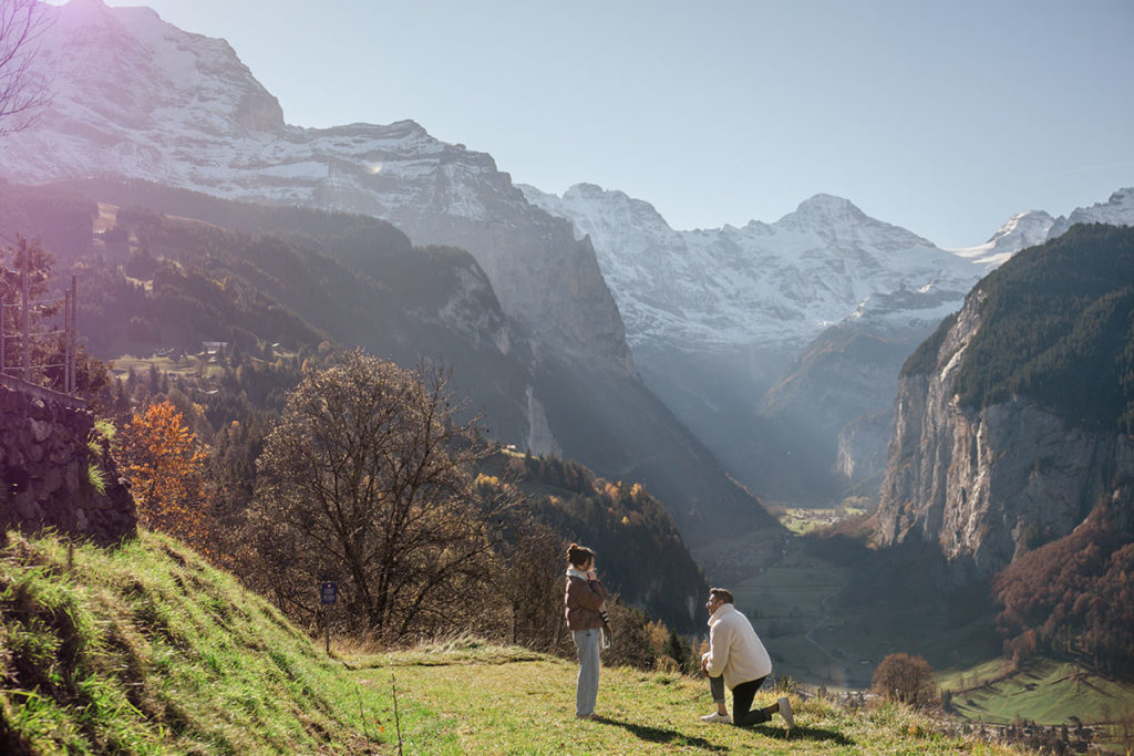 A person makes a surprise proposal on one knee to another person on a grassy hill, with mountains and the Lauterbrunnen valley in the background.