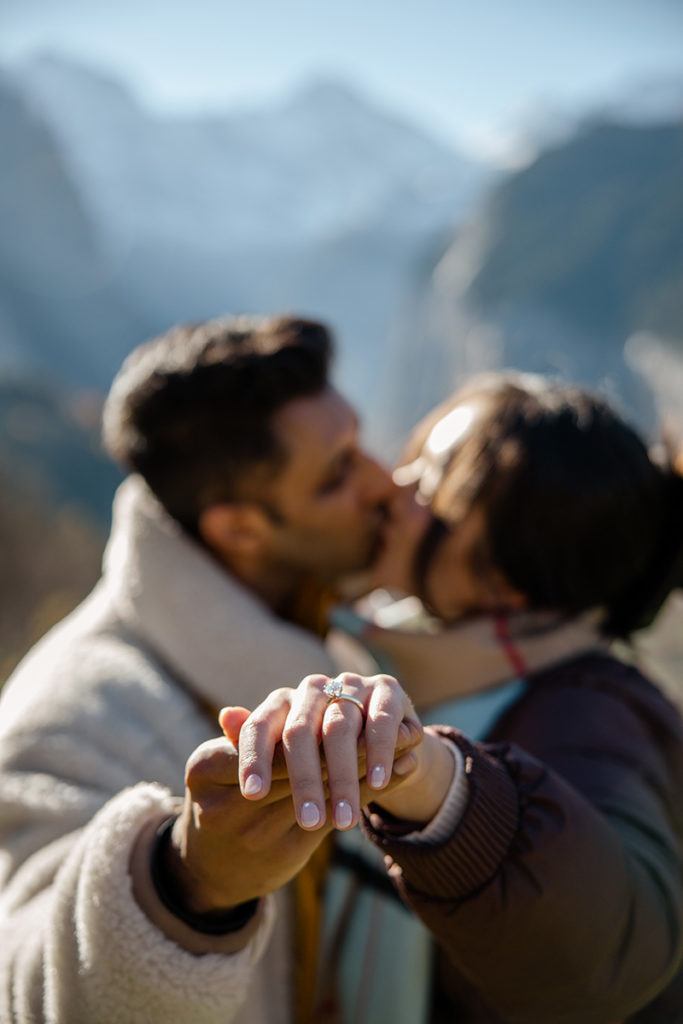 A couple kisses outdoors with mountains in the background, while one person holds out their hand to display an engagement ring in focus after a surprise proposal in Wengen, Switzerland.