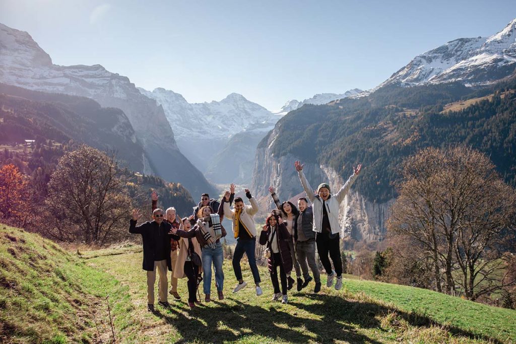 A group of people pose and jump for a photo on a grassy hill in Wengen with mountains and a clear sky in the background, during a surprise proposal in Switzerland