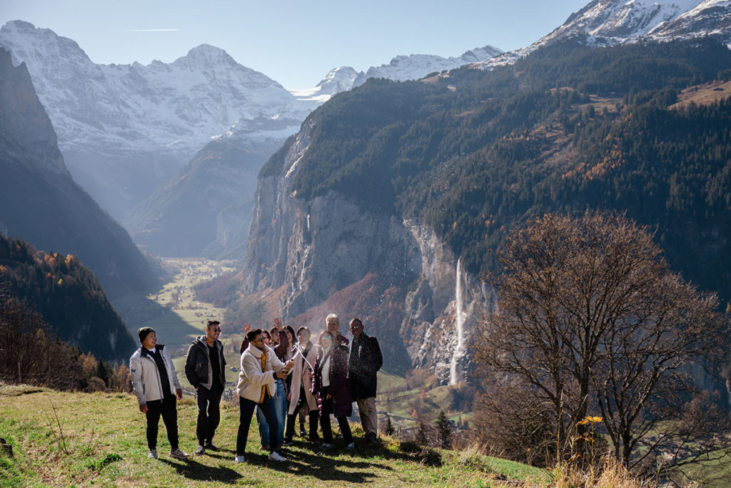 A group of people stands on a Wengen hillside taking a selfie, capturing the moment of a secret proposal, with mountains, cliffs, the waterfall and the Lauterbrunnen Valley in the background under a clear sky.