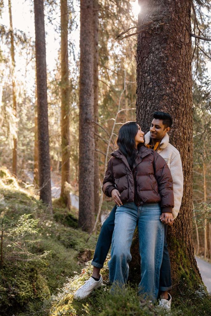 A couple stands close together by a tree in a sunlit forest in Wenden, both wearing casual jackets and jeans, capturing the magic of a Switzerland proposal.