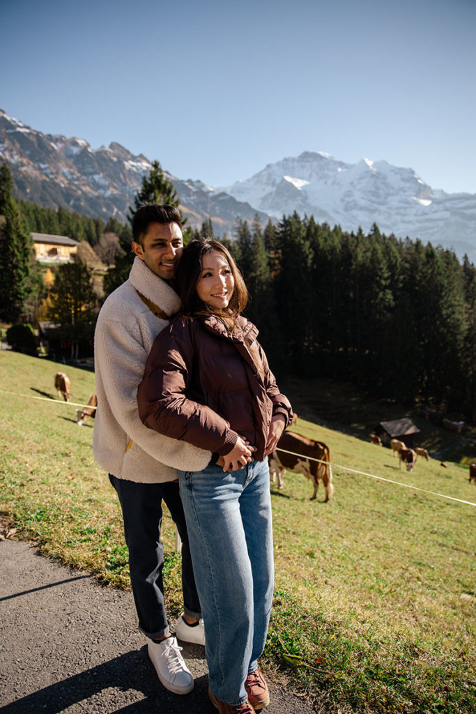 A couple stands together on a grassy hillside with cows grazing nearby and snow-capped mountains in the background, capturing the magic of a Wengen wedding proposal.