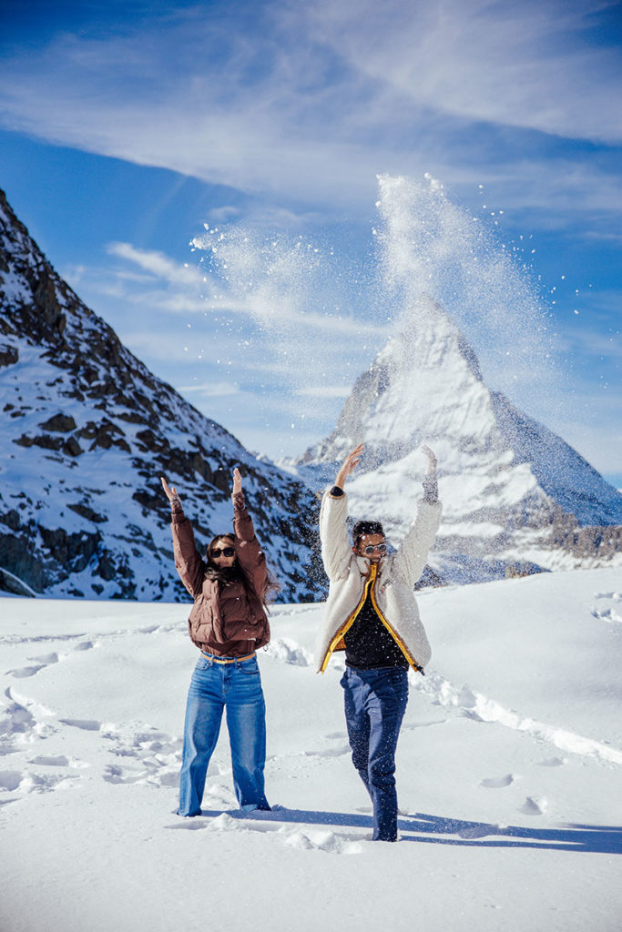 Two people stand in the snow with the Matterhorn in the background, throwing snow into the air on a sunny day during a destination Engagement shoot in Zermatt