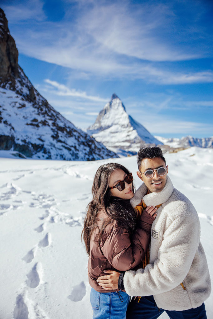 A couple in winter jackets poses on snowy ground with sunglasses, the Matterhorn and a clear sky in the background, during their destination Engagement shoot in Zermatt