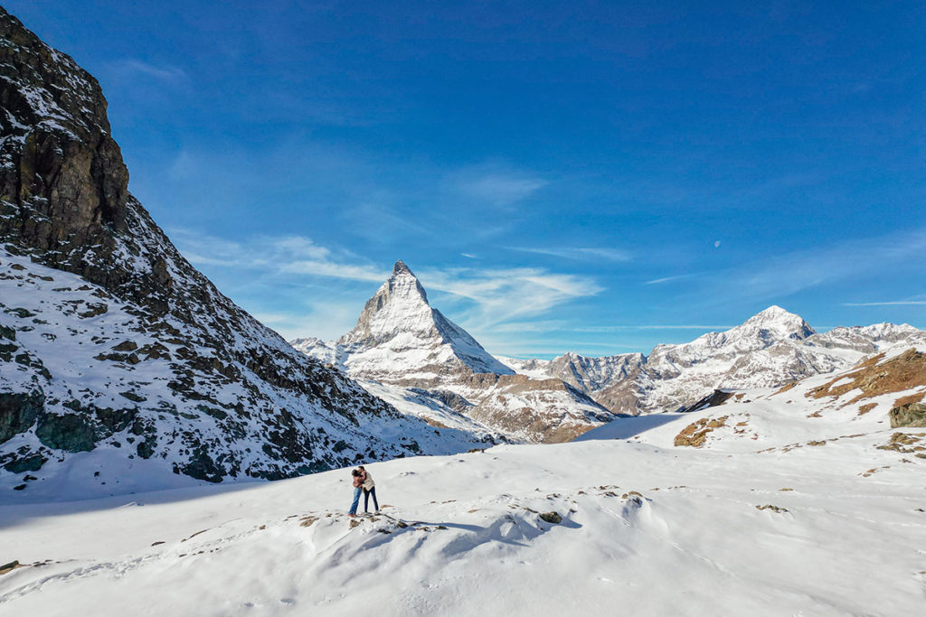 A couple stands on a snowy mountain slope, during their destination Engagement shoot in Zermatt with the Matterhorn peak visible in the background under a clear blue sky.