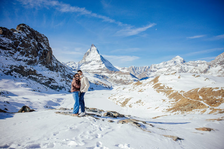 A couple stand embracing on a snowy mountain, the Matterhorn and clear blue sky behind them - a magical moment just during their destination engagement shoot in Zermatt