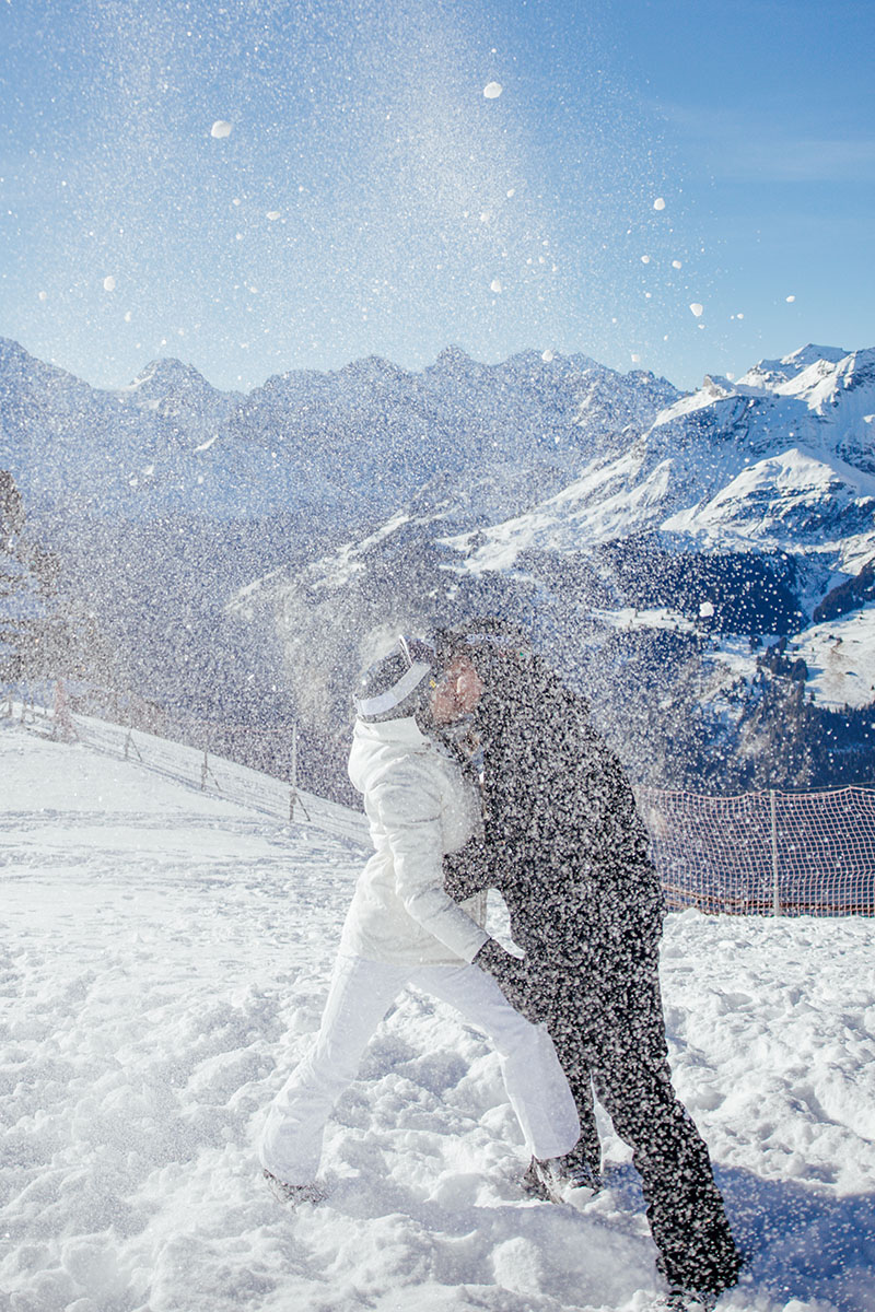 Two people in winter clothing playfully throw snow at each other on a snowy mountain, during their Proposal shoot with stunning snow-covered peaks and a clear blue sky in the background.